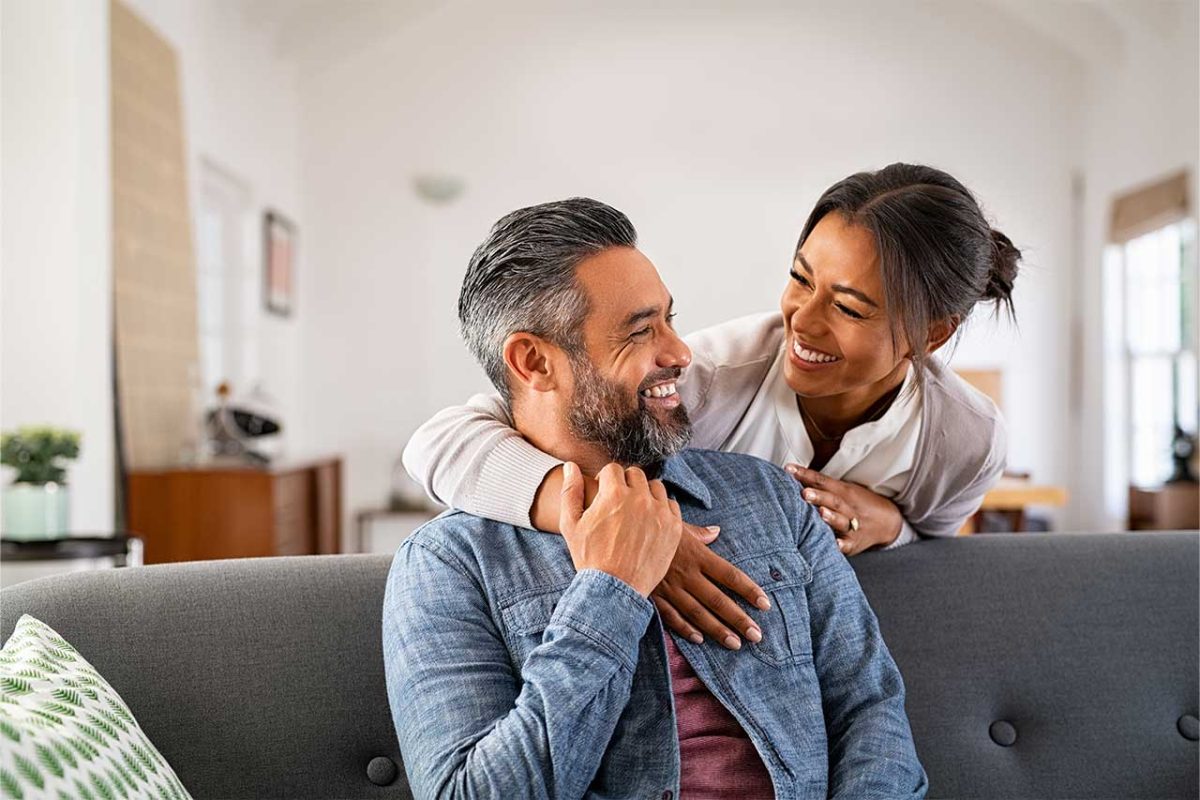 man and woman smiling with a woman's arm around his neck on a couch